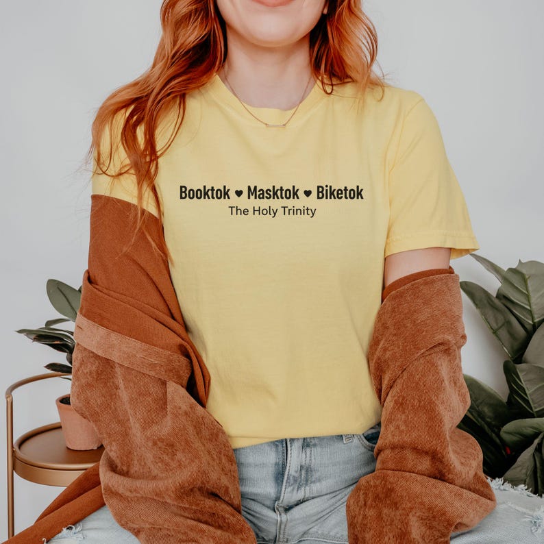 May include: Yellow t-shirt with the text "Booktok Masktok Biketok The Holy Trinity" in black. The person is wearing a brown jacket over the shirt and blue jeans. The background includes a plant and a small table.
