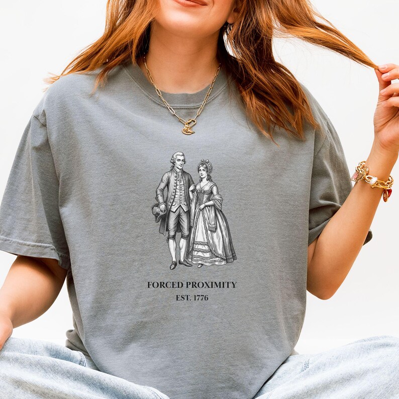 May include: Gray t-shirt featuring a black and white illustration of a couple in historical attire. The text "FORCED PROXIMITY EST. 1776" is printed below the image. The shirt has a relaxed fit and a crew neck.
