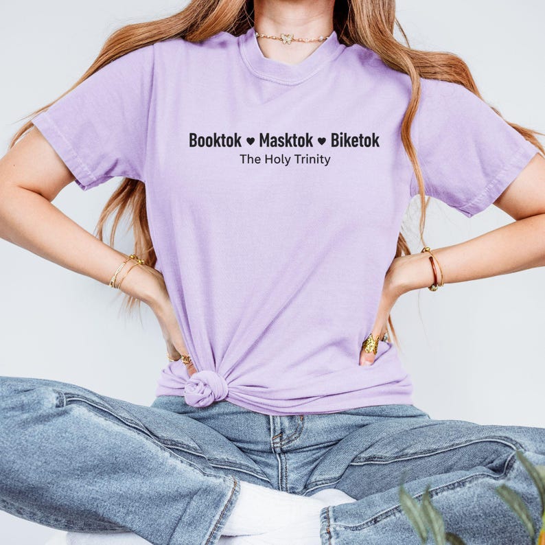 Pu&ograve; includere: Una maglietta lilla con la scritta "Booktok &hearts; Masktok &hearts; Biketok" sopra "The Holy Trinity" in nero. La maglietta &egrave; annodata in vita e abbinata a jeans blu. La modella indossa braccialetti e una collana dorati.