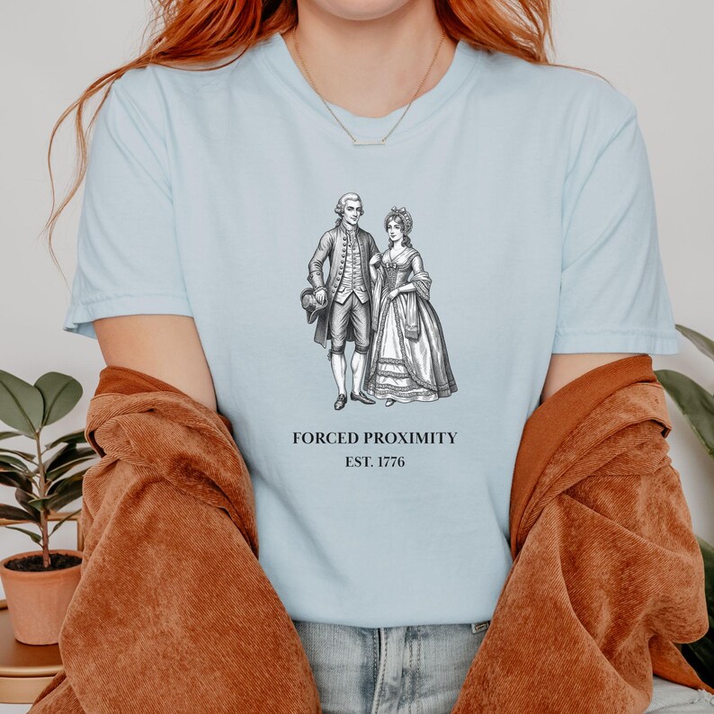 May include: Light blue t-shirt featuring a black and white illustration of a couple in historical attire. The text "FORCED PROXIMITY EST. 1776" is printed below the image. The shirt is a crew neck style.