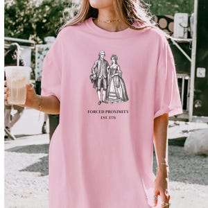 May include: A light pink t-shirt featuring a black and white illustration of a couple in historical attire. The text "FORCED PROXIMITY EST. 1776" is printed below the image. The shirt is a casual, oversized style.
