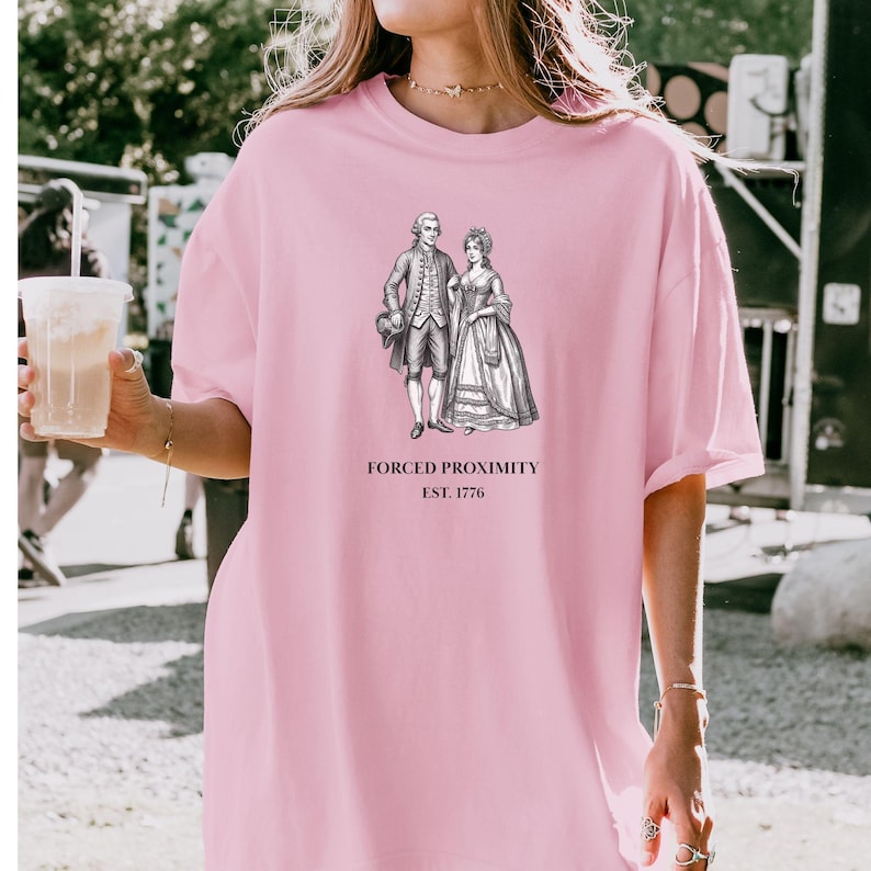 May include: A light pink t-shirt featuring a black and white illustration of a couple in historical attire. The text "FORCED PROXIMITY EST. 1776" is printed below the image. The shirt is a casual, oversized style.