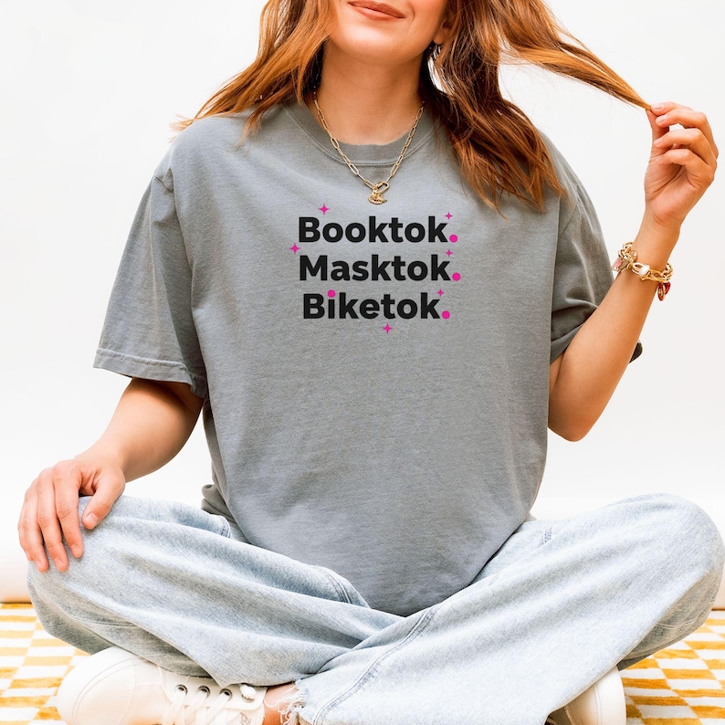May include: A gray t-shirt with the words "Booktok, Masktok, Biketok" in black text with pink star accents. The shirt is a casual style, suitable for everyday wear.