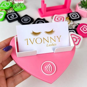 May include: A heart-shaped pink display stand with a white card holder featuring the gold text IVONNY Lashes and a gold eyelash graphic. The stand includes a white circular NFC icon and is surrounded by various social media logo keychains.