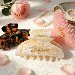 Könnte beinhalten: Drei Haarspangen sind abgebildet. Eine ist schildpattfarben mit "Eleni" in goldener Schrift, eine weitere cremefarben mit "Elyssia" und die dritte rosa mit "Stef" in goldener Schrift. Eine rosa Rose, Blütenblätter und eine Perlenkette sind ebenfalls vorhanden.