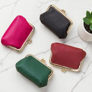 Puede incluir: Cuatro pequeños monederos en colores vibrantes: fucsia, negro, burdeos y verde esmeralda. Cada monedero tiene un marco de metal dorado con un cierre de boquilla. Los monederos están hechos de cuero texturizado y están colocados sobre una superficie de mármol blanco.