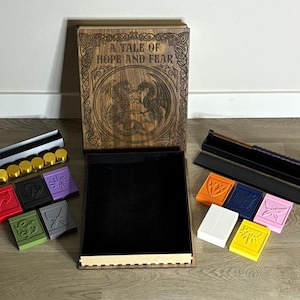 Peut inclure: Coffret en bois avec le texte "A TALE OF HOPE AND FEAR" et un motif de dragon. Comprend diverses pièces de jeu colorées, des supports à dés et un intérieur noir. L'ensemble est conçu pour les jeux de société.