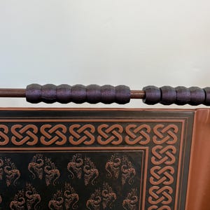 May include: A decorative wooden banner with a dark brown frame and a repeating Celtic knot design. The banner has a dark background with a repeating pattern of stylized dragons. The banner is held by a brown rod with padded grips.