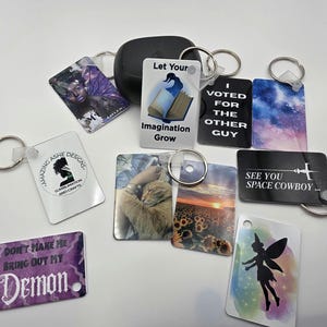 May include: A collection of keychains with various designs. One keychain features a woman with purple wings, another has the text "Let Your Imagination Grow", and another says "I Voted For The Other Guy". Other keychains have images of a cat, sunflowers, and a fairy.