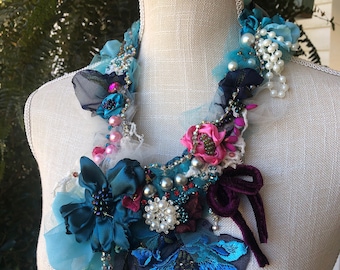 Puede incluir: Un collar floral azul, rosa y blanco con detalles de perlas y un lazo de terciopelo. El collar está hecho de tela, cuentas y cinta.