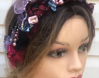 Puede incluir: Una diadema floral de color burdeos, negro y morado con pedrería y perlas. La diadema está hecha de tela y encaje.