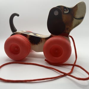 May include: Vintage pull-along toy dog with red wheels and a red pull string. The dog has a brown and white spotted body, a black tail, and a brown and black head. The toy is designed to be pulled along the floor.