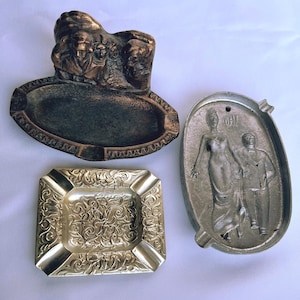 May include: Three vintage ashtrays. One is bronze-toned with a sculpted relief of figures. Another is silver-toned, rectangular, and features an ornate floral design. The third is oval-shaped with a couple and the word "OH!"