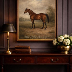 May include: An oil painting of a brown horse in a field, framed and displayed above a dark wood console table. The table has a brass lamp, two leather-bound books, and a gold bowl of white flowers. The wall is a dark brown colour.