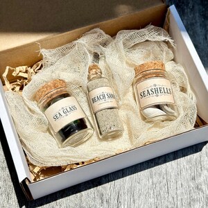 May include: Three clear glass jars with cork stoppers nestled in beige cheesecloth inside a white box. The jars have labels reading SEA GLASS, BEACH SAND, and SEASHELLS. The jars contain dark sea glass, fine sand, and small white seashells respectively.
