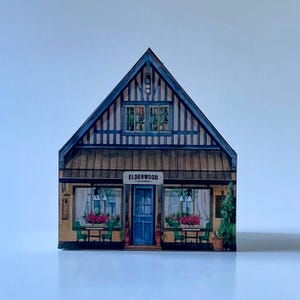 May include: A decorative wooden block shaped like a house facade with a blue and tan exterior. The front features a sign reading ELDERWOOD KITCHEN, a central blue door, large windows with white curtains, green chairs, and pink floral accents.