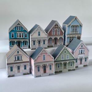 May include: A collection of eight miniature house models featuring various architectural details, including gabled roofs, porches, and windows. The houses are painted in diverse colors such as blue, cream, pink, green, and brown, arranged in two rows.