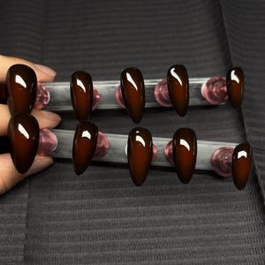 Puede incluir: Un conjunto de diez uñas postizas brillantes en forma de almendra, en un tono marrón oscuro y rico. Las uñas están dispuestas sobre soportes transparentes con detalles rosas, mostrando su superficie lisa y reflectante. Las uñas son de un color oscuro y sólido.
