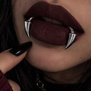 Könnte beinhalten: Ein Paar silberfarbene, gebogene, fangzahnförmige Lippenpiercings an den Mundwinkeln. Der Schmuck hat eine glatte, metallische Oberfläche mit spitz zulaufenden Enden, die nach unten zeigen und einen Kontrast zum dunklen, matten Lippenstift bilden.