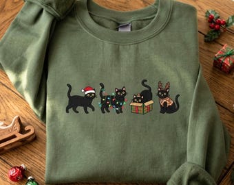 May include: An olive green crewneck sweatshirt featuring a graphic print of four black cats. One cat wears a Santa hat, one is tangled in colorful string lights, one sits inside a plaid gift box, and one wears reindeer antlers and a sweater.
