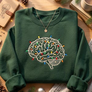 May include: A dark green crewneck sweatshirt featuring an embroidered graphic of a human brain wrapped in colorful Christmas lights. A delicate gold-tone necklace with a small circular pendant rests on the neckline of the garment.
