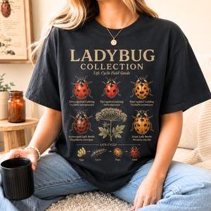 May include: A black t-shirt featuring a graphic print titled LADYBUG COLLECTION Life Cycle Field Guide. The design includes illustrations of six ladybug species, a plant, and a life cycle diagram with labels for eggs, larva, pupa, and adult.