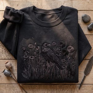 May include: A black crewneck sweatshirt featuring detailed tonal embroidery of a raven perched among various wildflowers and grasses. The design is centered on the front of the garment, showcasing intricate stitching that creates texture and depth.
