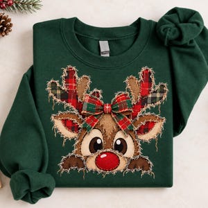 May include: A dark green crewneck sweatshirt featuring a central applique of a reindeer face. The reindeer has a bright red nose, brown fur, and plaid fabric antlers and ears with frayed edges. A plaid bow sits between the antlers.