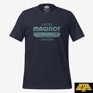 May include: Navy blue t-shirt with the text "USCSS MAGINOT" in teal. Below the text is a teal graphic and the number "294068". The shirt has short sleeves and a crew neck. The brand logo "INTER GALACTIC FIT" is in the bottom right corner.
