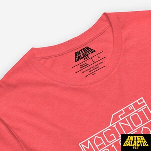 Peut inclure: T-shirt corail avec le texte blanc "MAGINOT" et le logo "INTER GALACTIC FIT". L'&eacute;tiquette indique qu'il est fabriqu&eacute; au Nicaragua et est un m&eacute;lange de coton et de polyester.