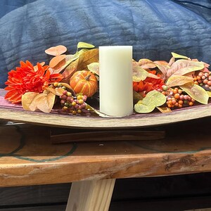 Può includere: Un vassoio decorativo in legno con una composizione a tema autunnale. Presenta una candela color crema, fiori arancioni, piccole zucche e bacche colorate. Il vassoio è posto su un tavolo di legno, creando un'atmosfera calda.