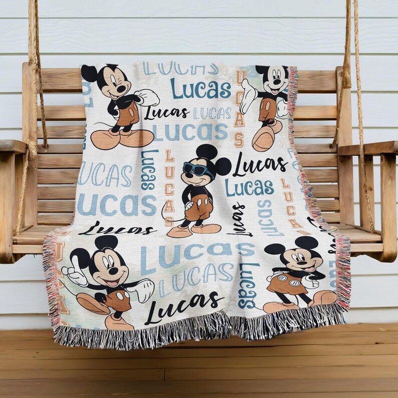 A Mickey Mouse-themed throw blanket with the name "Lucas" printed on it, hanging on a wooden bench.