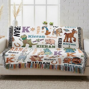 a colorful throw blanket with various cartoon animal characters and the letters "Kieran" printed on it. The blanket is placed on a couch, and there are some potted plants in the background.