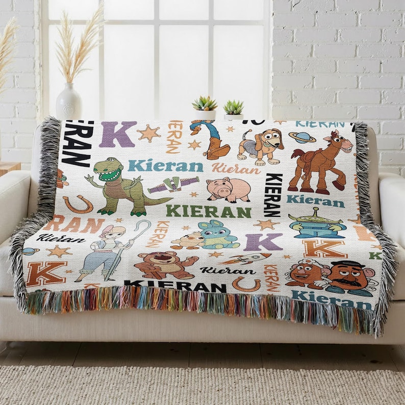 a colorful throw blanket with various cartoon animal characters and the letters "Kieran" printed on it. The blanket is placed on a couch, and there are some potted plants in the background.