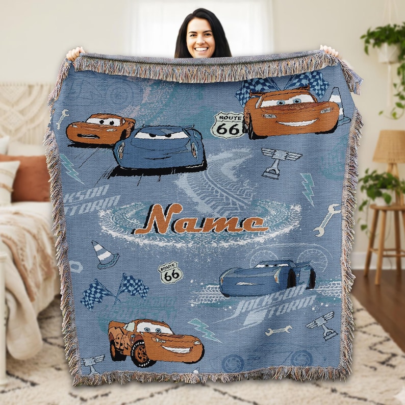 A woman is holding up a blue blanket with a design featuring cartoon cars and the word "Name" printed on it.