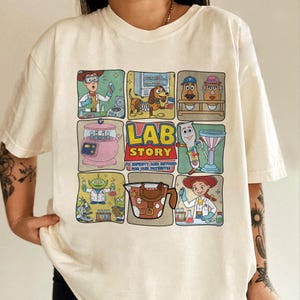 May include: Cream-colored t-shirt featuring a colorful graphic design. The design includes cartoon characters in a lab setting with the words "LAB STORY" and the phrase "TO INFINITY AND BEYOND FOR OUR PATIENTS!"