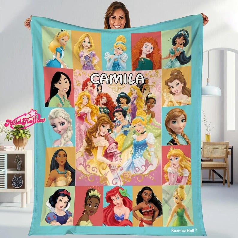 A woman is holding up a colorful blanket featuring various Disney princess characters, including Snow White, Rapunzel, and other popular Disney princesses.