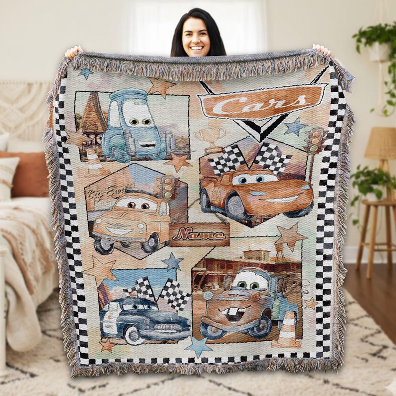 A woman is holding up a colorful blanket featuring various cartoon cars, including the Cars characters from the Disney animated film.