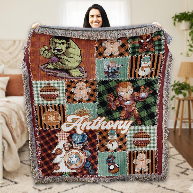 A woman is holding up a colorful, patchwork blanket featuring various cartoon characters and designs, including the Hulk, Spider-Man, and other popular superheroes.