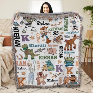A woman is holding up a colorful, embroidered blanket featuring various characters from the Toy Story franchise, including Woody, Buzz Lightyear, and other beloved characters.