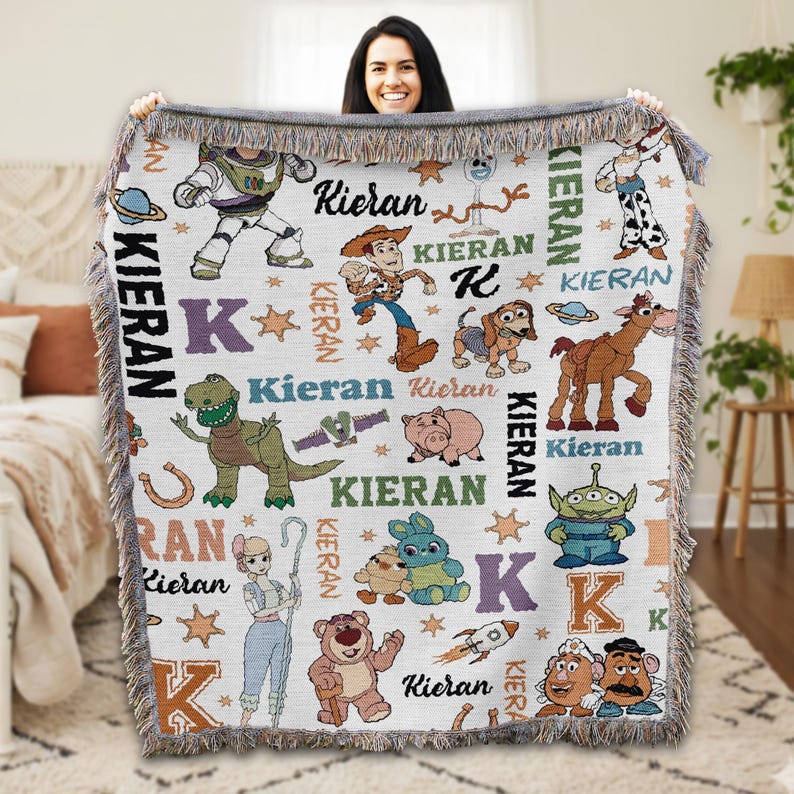 A woman is holding up a colorful, embroidered blanket featuring various characters from the Toy Story franchise, including Woody, Buzz Lightyear, and other beloved characters.