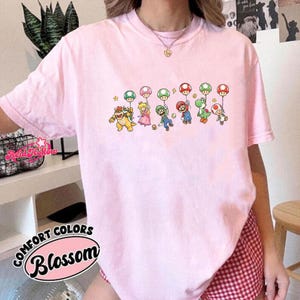 May include: A light pink short-sleeve t-shirt featuring a graphic print of various video game characters, including Bowser, Princess Peach, Luigi, Mario, Yoshi, and Toad, each holding a colorful mushroom balloon. The shirt has a crew neckline.