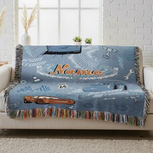 A blue throw blanket with a Cars-themed design, featuring a race car, tires, and other car-related elements, is draped over a white couch.