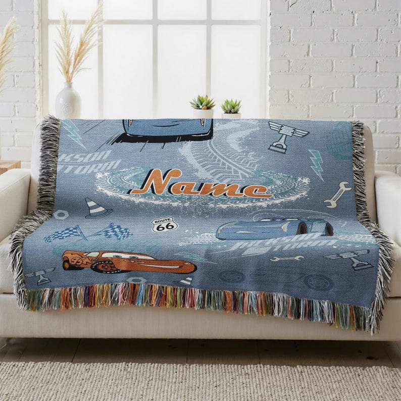 A blue throw blanket with a Cars-themed design, featuring a race car, tires, and other car-related elements, is draped over a white couch.