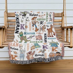 A colorful throw blanket with various cartoon characters and the name "Kieran" printed on it, hanging on a wooden bench.