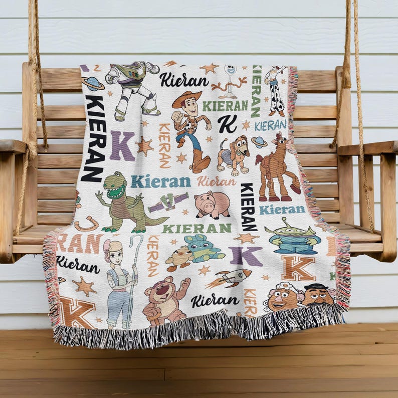 A colorful throw blanket with various cartoon characters and the name "Kieran" printed on it, hanging on a wooden bench.