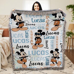 A woman is holding up a Mickey Mouse-themed blanket with the name "Lucas" written on it.