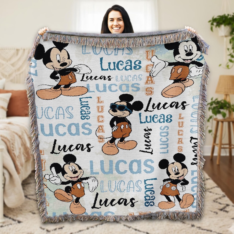 A woman is holding up a Mickey Mouse-themed blanket with the name "Lucas" written on it.