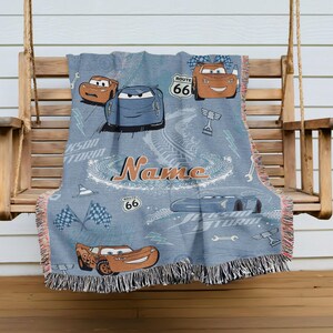 A blue blanket with a Cars-themed design, featuring various cars and racing elements, is hanging on a wooden bench.