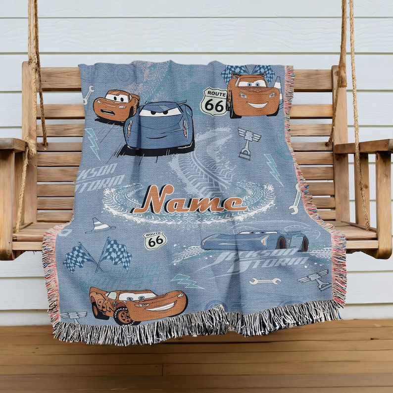A blue blanket with a Cars-themed design, featuring various cars and racing elements, is hanging on a wooden bench.